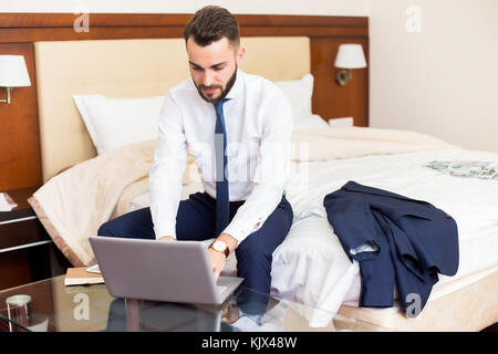 Portrait of handsome bearded businessman using laptop working in hotel room sitting on bed and enjoying business travel, copy space Stock Photo