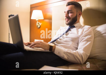 Portrait of handsome bearded businessman using laptop in bed enjoying hotel stay during business travel Stock Photo