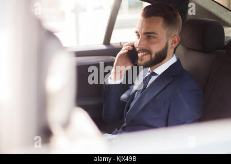 Portrait of handsome bearded businessman speaking by phone sitting in back seat of luxury car and smiling Stock Photo