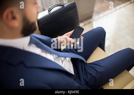 High angle close up of unrecognizable businessman using smartphone waiting in lobby with leather case Stock Photo