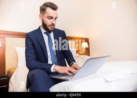 Portrait of handsome bearded businessman using laptop sitting on bed in comfortable hotel room during business travel, copy space Stock Photo