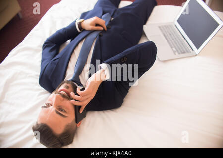 Above view of happy handsome businessman speaking by phone and smiling laying on hotel bed next to laptop, enjoying business travel, copy space Stock Photo