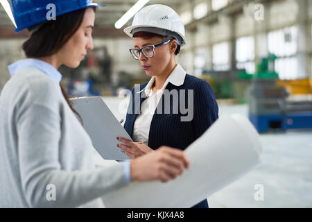 Serious busy lady in hardhat working in factory and making notes while holding examination together with engineer Stock Photo