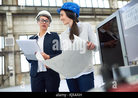 Confident female technologist in hardhat examining document while engineer showing blueprint in factory Stock Photo