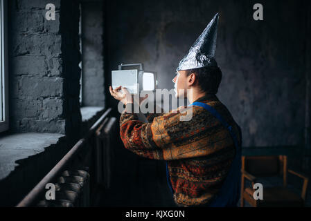 Paranoid man in tinfoil cap watch TV, mind protection from telepathy, paranoia concept. UFO phobia Stock Photo