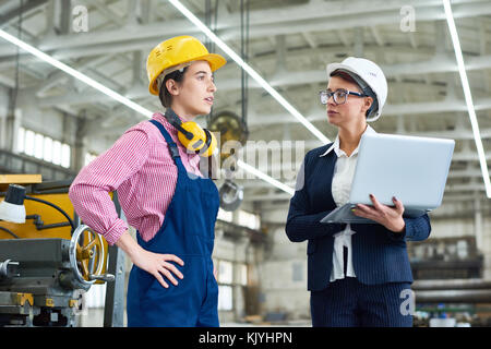 Serious confident female engineer in formal suit talking to manual worker with protective ear muffs on neck working in factory workshop Stock Photo