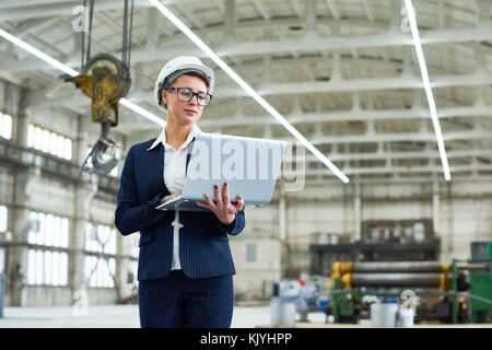 Pensive attractive businesswoman running industrial factory and visiting production workshop using portable computer while standing alone at construct Stock Photo