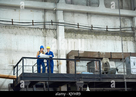 Busy confident female factory workers analyzing blueprint while working in old factory and standing on industrial bridge Stock Photo