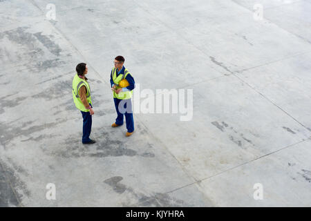 Directly above view of serious female construction workers in green waistcoat standing on open space in factory and discussing building structure Stock Photo