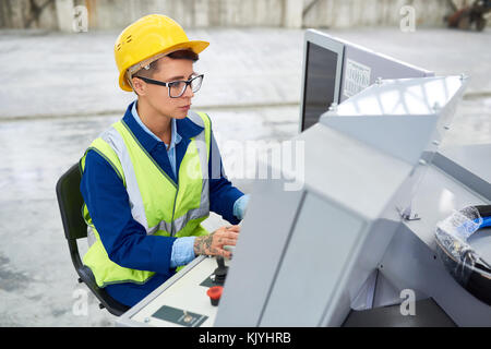 Concentrated factory employee in green waistcoat using technical machine while working on operator dashboard at the plant Stock Photo