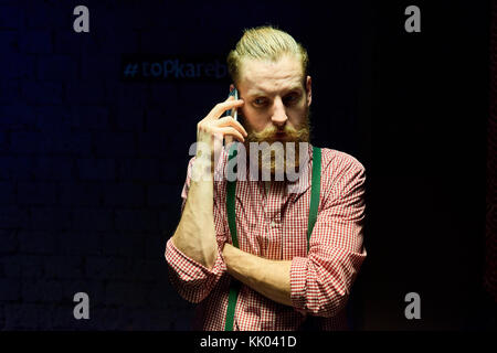 Portrait of handsome bearded hipster posing speaking by phone against black background drowning in harsh contrasted spot light from above Stock Photo