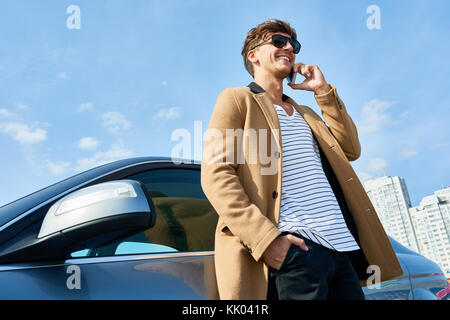 Low angle portrait of handsome young man speaking by phone standing in sunlight leaning on car outdoors against clear blue sky, copy space Stock Photo