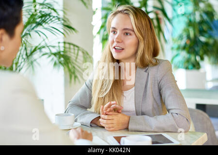 Portrait of two modern young women discussing work sitting at table in cafe during business meeting, focus on young blond trainee talking enthusiastic Stock Photo