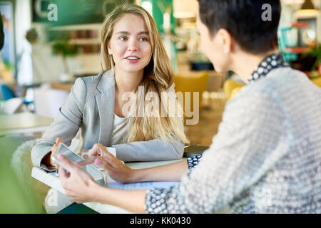 Portrait of smiling young businesswoman showing digital tablet to colleague explaining project during business meeting in cafe Stock Photo