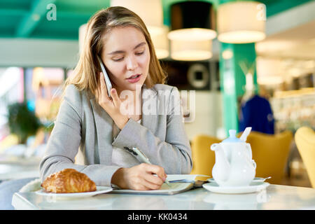 Portrait of pretty young businesswoman speaking by phone and making notes while working in cafe during lunch break Stock Photo