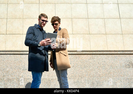 Portrait of two handsome young businessmen using digital tablet outdoors standing against background of concrete wall, copy space Stock Photo