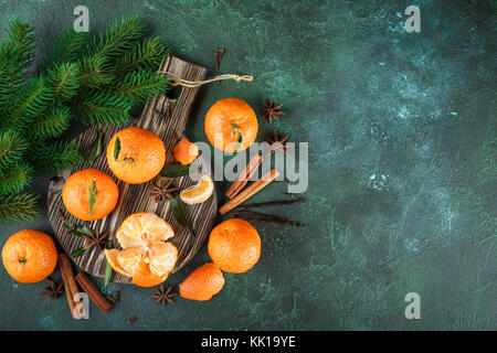 Fresh mandarins with leaves and spices - cinnamon, anise and cloves on a dark cutting board on a dark green background. Top view Stock Photo