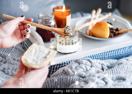 Female hands smearing cream cheese on fresh bread. Breakfast in bed, a tray with cheese, grissini, jam and aromatic candle. Christmas morning. Honeymo Stock Photo
