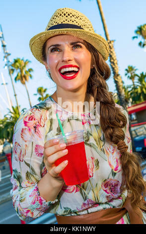 Summertime at colorful Barcelona. smiling elegant tourist woman in long dress and straw hat on embankment in Barcelona, Spain with bright red beverage Stock Photo