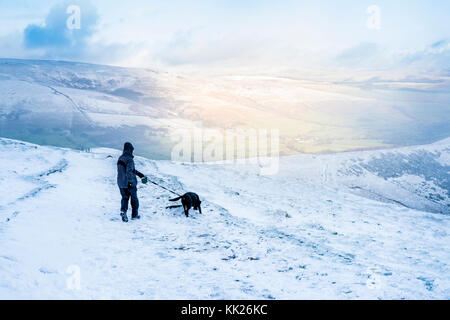 Dog walking in the snow. Hiker with dog hiking with sunlight shining on a hill across the valley. Great Ridge, Derbyshire, Peak District, England, UK Stock Photo