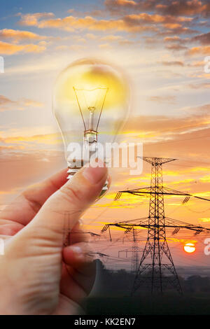 electricity industry- hight voltage energy - hand holding a glowing bulb with power station in the background, sunset and the electrical poles Stock Photo