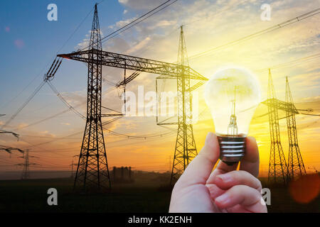 electricity industry- hight voltage energy - hand holding a glowing bulb with power station in the background, sunset and the electrical poles Stock Photo