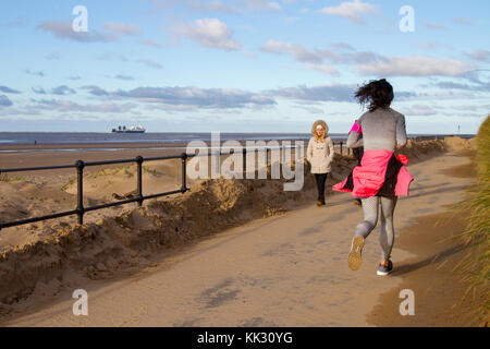 Woman running on the seafront promenade at Crosby, Merseyside.  UK Weather. November, 2017. Bright and sunny on the west coast of the Mersey Estuary. This coastal area around Crosby beach, and Crosby Marine Park stretches from Waterloo to the estuary of the River Alt at Hightown. The beach and dunes is currently home to 100 metal human sculptures Antony Gormley's Another Place. There are dangerous quicksands so visitors to the shoreline are advised to keep within 50 metres of the promenade. Credit; MediaWorldImages/AlamyLiveNews Stock Photo