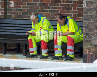 Pair of male workmen sitting taking a break while using mobile phones. Stock Photo