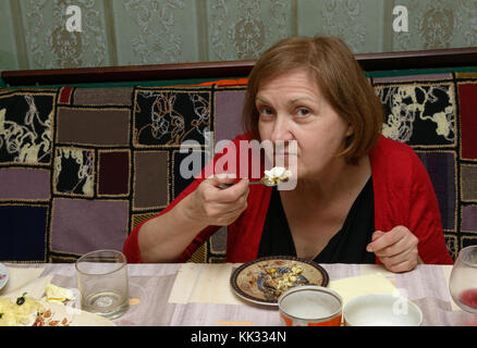 Close-up portrait of elderly woman inn red sweater that is eating with funny facial expression. Stock Photo