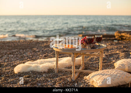 Romantic outdoor dinner with wine and cheese by seaside. Two glasses of wine, grapes, dried fruits and cheese placed on small round table, with white  Stock Photo