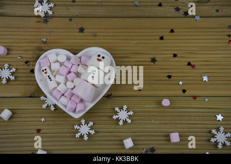 romantic winter season photography image with white love heart shape dish filled with marshmallows and some shaped as snowman with iced on smile Stock Photo