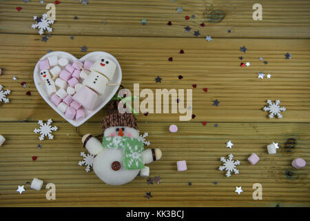 romantic winter season photography image with white love heart shape dish filled with marshmallows and some shaped as snowman with iced on smile Stock Photo
