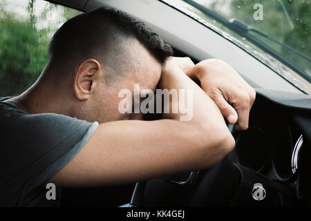 closeup of a young caucasian man sitting at the driver seat of a car reclining his head on the steering wheel, in a rainy day Stock Photo