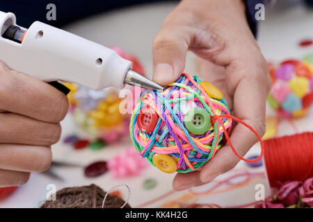 closeup of a young man making a handmade christmas ball using a hot glue gun with strings and buttons of different colors, and a pile of different hab Stock Photo