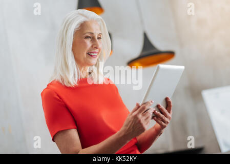 Innovative device. Cheerful positive elderly woman looking at the tablet screen and smiling while using modern technologies Stock Photo