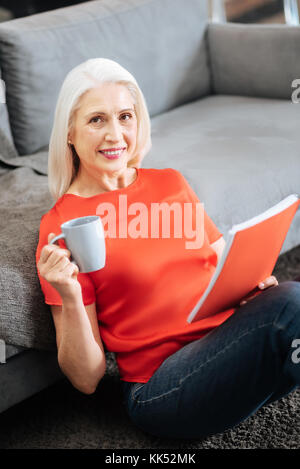 Delicious drink. Delighted happy positive woman having tea and reading her notebook while looking at you Stock Photo