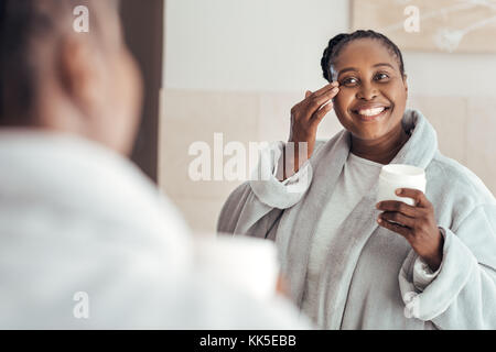 Smiling young African woman putting on face cream at home Stock Photo