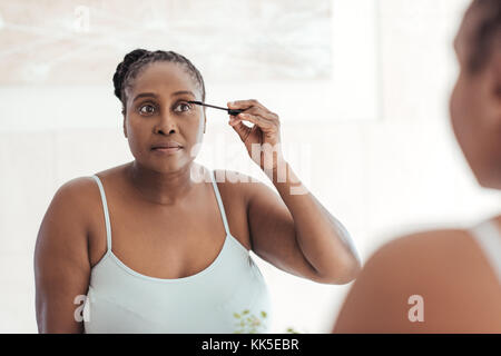 African woman applying mascara in a bathroom mirror at home Stock Photo