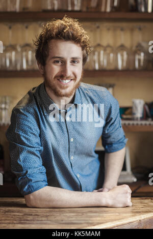 Young barman leaning on wooden bar counter and smiling at camera, shelves with glasses on background Stock Photo