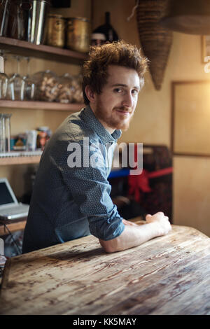 Young barman leaning on wooden bar counter and smiling at camera, shelves with glasses on background Stock Photo