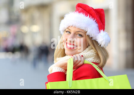 Portrait of a happy girl wearing santa claus hat shopping in christmas looking at you outdoors on the street Stock Photo