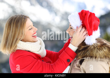 Side view portrait of two friends joking in christmas holidays with a santa claus hat with a snowy mountain in the background Stock Photo