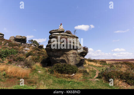 A young man sits on top of a rock formation at Brimham rocks in North Yorkshire on a summers day. Stock Photo