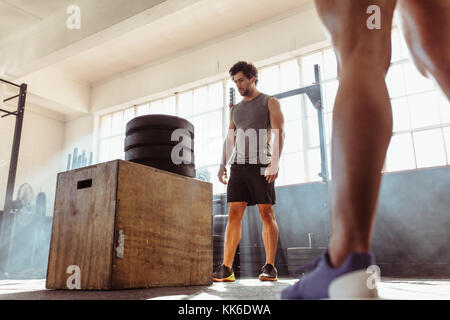 Determined fit man doing box jumping in health club. Muscular male athlete standing by wooden box with extra height made by pile of weights. Stock Photo
