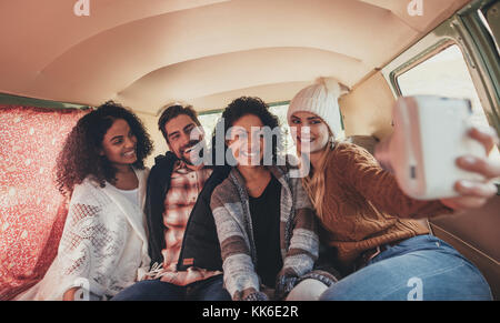 Friends sitting together inside van and taking selfie on road trip. Woman making selfie with friends using digital camera. Stock Photo