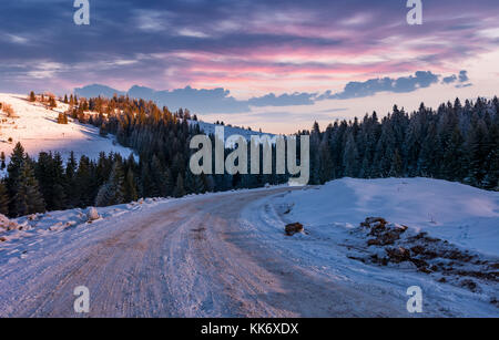 road through snowy hill side in to the spruce forest. gorgeous countryside landscape at winter dawn with magenta sky Stock Photo