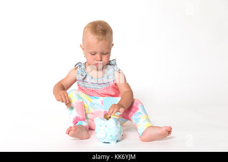 Cute toddler girl with a serious expression sitting on the floor is giving a coin to the saving piggy bank. All is on the white background. Horizontal Stock Photo