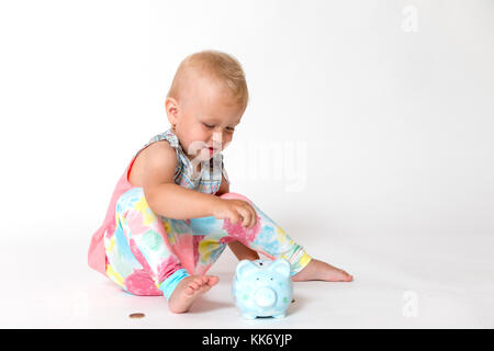 Smiling toddler girl sitting on the floor is giving one of the coins to the saving piggy bank. All is on the white background. Horizontally. Stock Photo