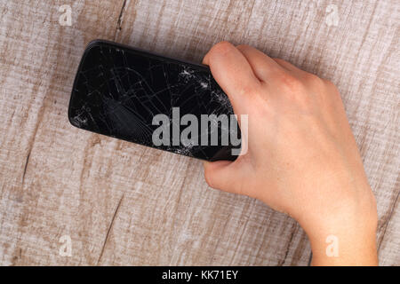 A smartphone with a broken screen in the girl's hand on a wooden table. Close-up. Studio lighting. Stock Photo