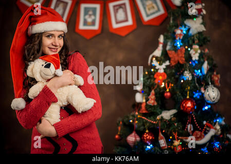 Lovely portrait of the charming smiling pregnant woman with santa calus hat holding the teddy bear toy at the blurred background of the decorated Christmas tree. Stock Photo